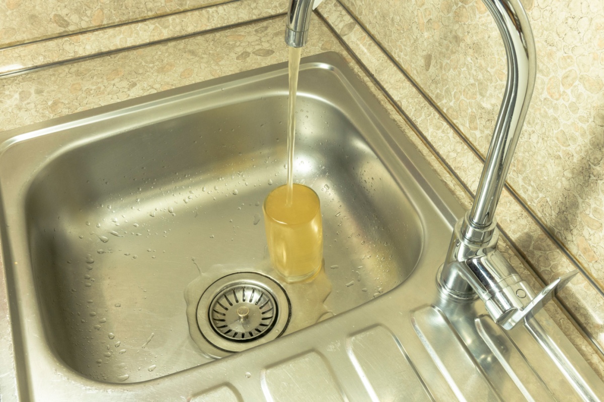 A yellowish water pours from a kitchen sink faucet into a cup.